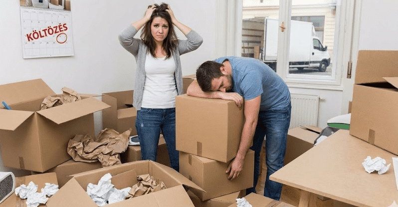 Egy stresszes pár feje fájva és kimerülten áll egy rendetlen, dobozokkal teli szobában egy budapesti költözés előtt, a falon egy "KÖLTÖZÉS" feliratú naptárral.
